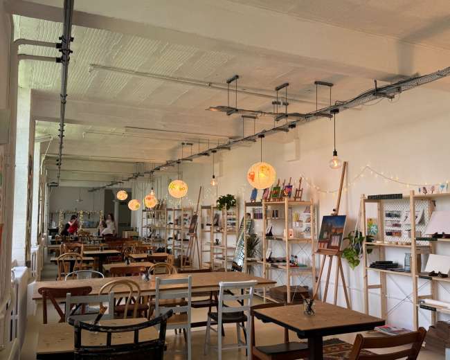 The image shows a modern café interior with wooden tables, hanging pendant lights, and decorative shelves displaying various items along the walls.