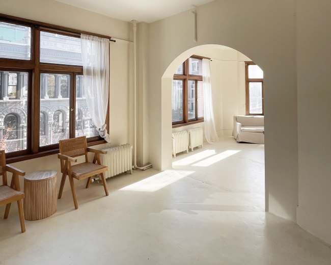 A well-lit, empty room with large windows, two wooden chairs, a round table, and an archway leading to another area.