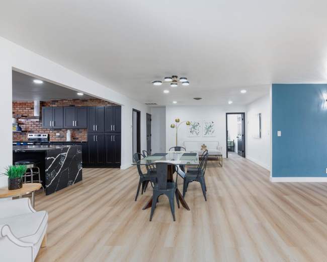 The image shows a spacious, modern dining area with a glass dining table and black chairs, adjacent to a kitchen with dark cabinetry and exposed brick walls.