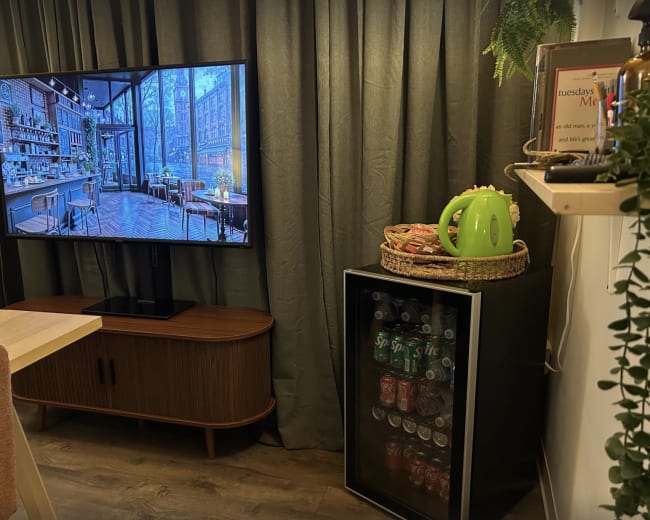 The image shows a living space with a large television displaying a café scene, a wooden sideboard, and a glass-fronted beverage fridge stocked with cans.