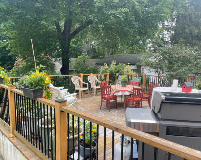A wooden deck features a table and chairs surrounded by potted plants and a grill, with trees and houses in the background.