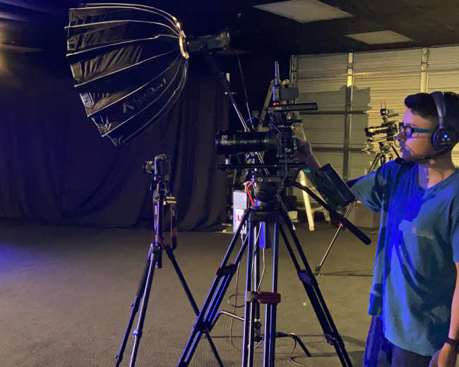 A person operates a camera and lighting equipment in a dimly lit studio setup.
