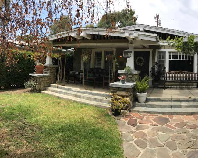 The image shows a single-story house with a wide porch, surrounded by a landscaped yard featuring stone pathways and various plants.