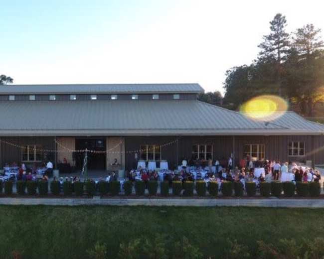 A large gathering of people is seated at tables arranged outside a metal building, with decorations and greenery lining the area.