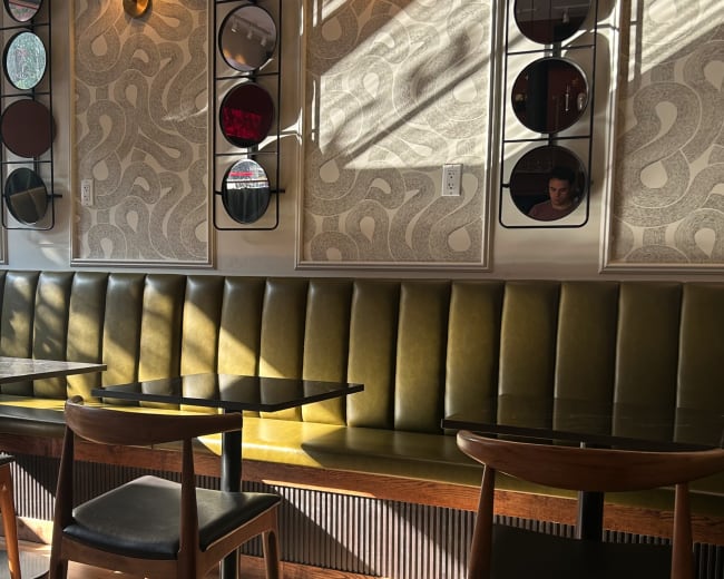 The image shows an interior of a café with green upholstered benches, wooden tables, and patterned wall decor, illuminated by sunlight streaming through the windows.