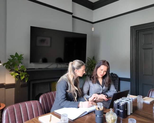 Two women are sitting at a conference table, collaborating on a laptop in a professional meeting space with a large television screen and decorative plants in the background.