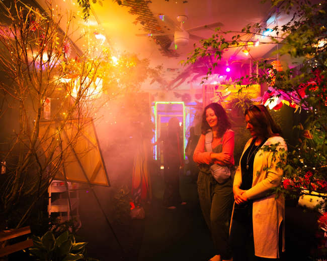 The image shows two women in a colorful, misty room filled with plants and vibrant lights, engaging in conversation.