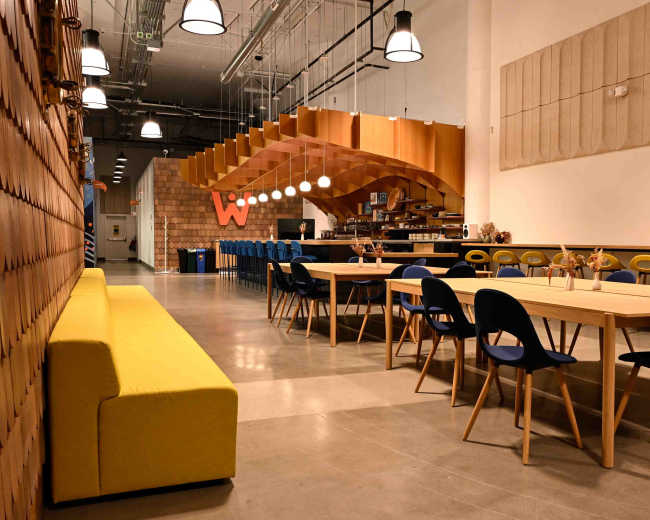 The image shows a modern, open dining area featuring a long wooden table with blue chairs, a yellow sofa along one wall, and wooden pendant lights above.
