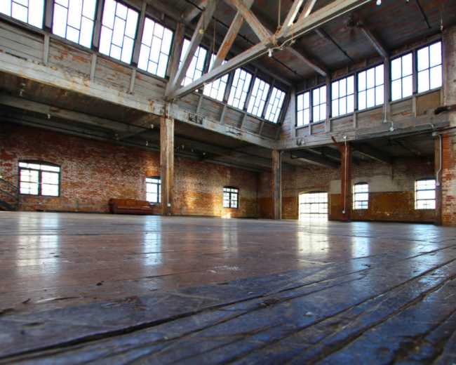 The image shows a spacious, industrial-style room with high ceilings, large windows, and exposed brick walls.