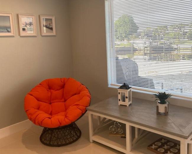 A bright orange chair is positioned next to a wooden table with decorative items in a sunlit room.