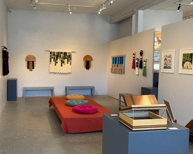 A spacious art gallery features colorful textile artworks on the walls, a red couch with cushions in the center, and a display stand on the right.