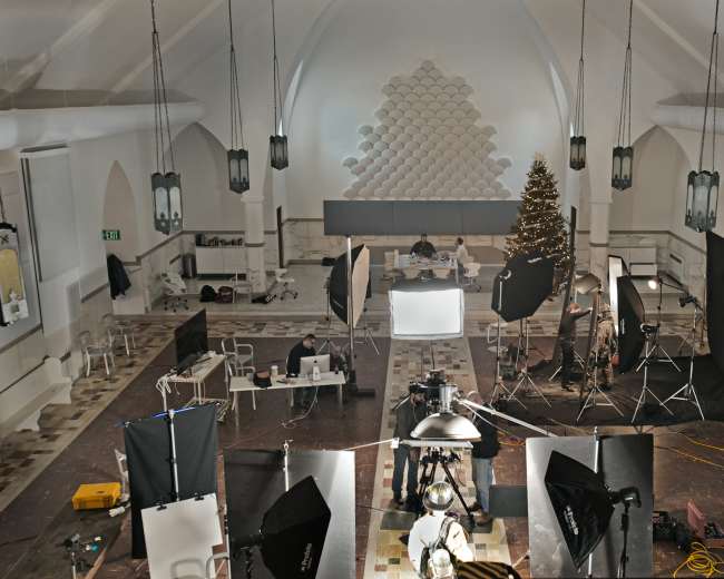 A large, well-lit studio space features multiple photography lights and equipment arranged around a central table with individuals working at computers, while a Christmas tree stands in the corner.