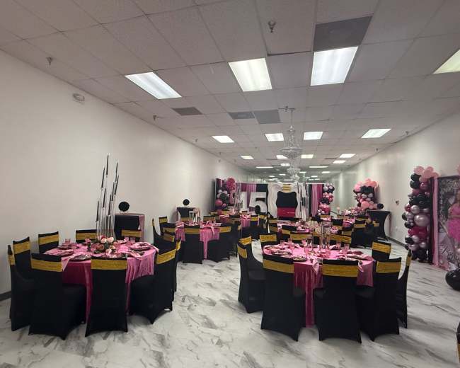The image shows a decorated event space with multiple round tables covered in pink tablecloths and black chairs, featuring centerpieces and balloon arrangements for a celebration.