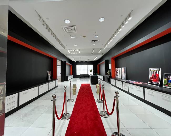 The image shows a modern exhibition space with a red carpet leading through a corridor flanked by black walls and white cabinets, featuring several framed photographs on display.