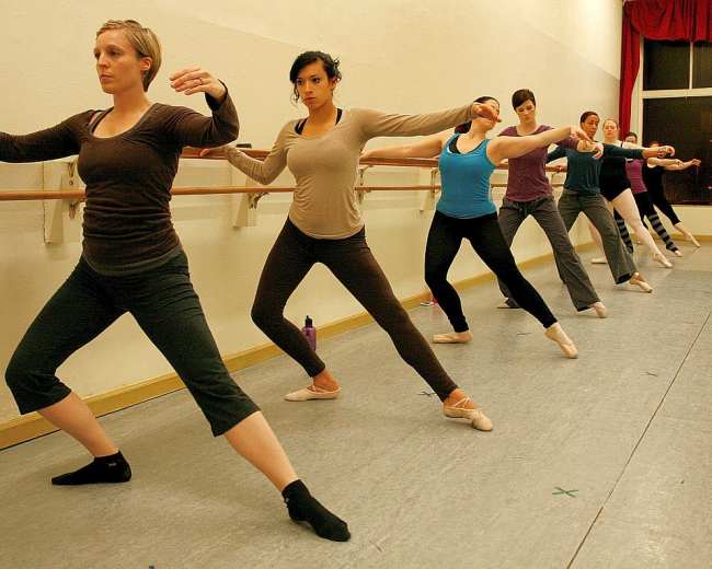A group of dancers performs a ballet exercise at a studio, with each dancer facing the same direction along the ballet barre.