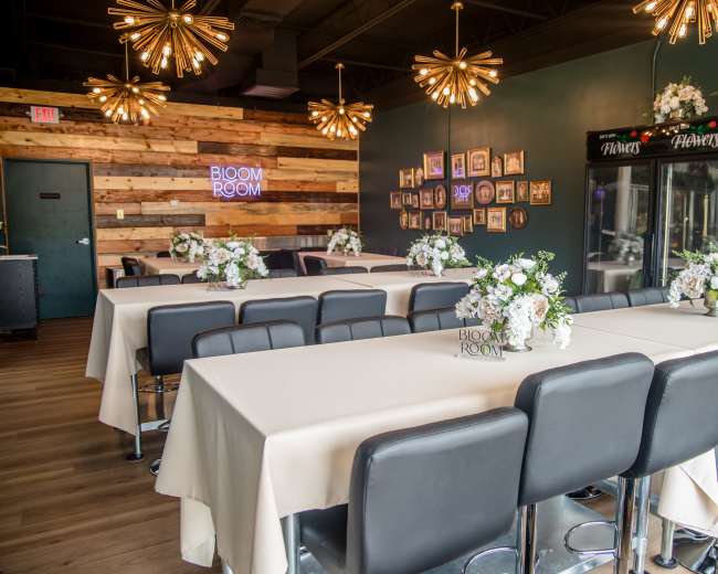 The image shows a stylish event space with multiple tables dressed in white tablecloths, decorated with flower arrangements, and a decorative wall featuring framed pictures and a neon sign that reads "BLOOM ROOM."