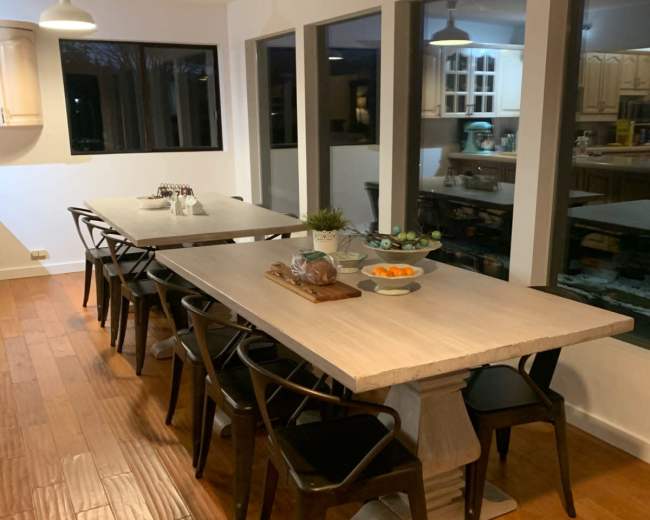 A long dining table with black chairs is set against a backdrop of large windows in a modern kitchen.