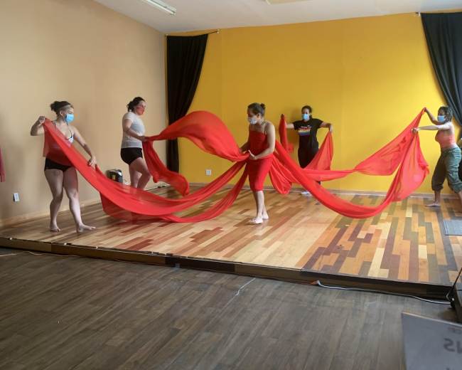 A group of dancers in leotards manipulate large red fabrics inside a dance studio with wooden flooring and yellow walls.