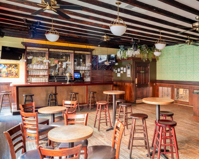 The image shows an interior view of a bar with wooden tables and chairs arranged around a polished wooden counter, illuminated by overhead lights.