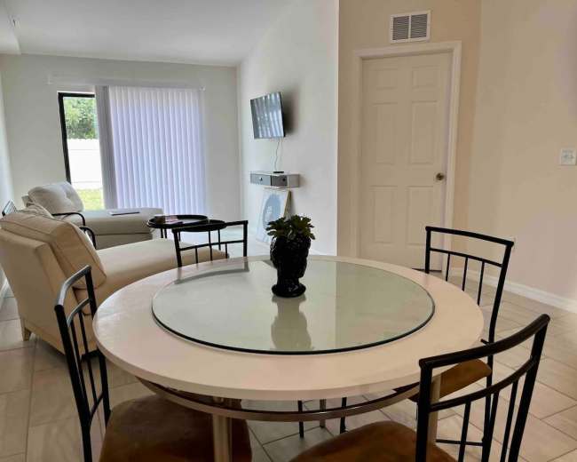 A round glass dining table with black chairs is positioned in a bright room featuring a sofa, a wall-mounted TV, and a door leading outside.