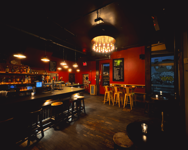 The image shows a dimly lit bar featuring a long counter with stools, a wall-mounted drink menu, and a chandelier, with graffiti visible through a window.