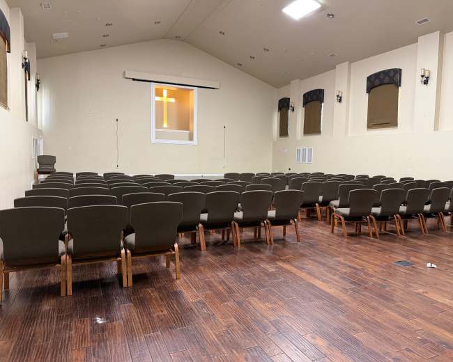 The image shows an empty auditorium with rows of chairs facing a front stage area, featuring a large window with a cross visible.