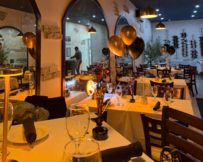 The image shows a restaurant interior decorated with black and gold balloons, featuring tables set with white tablecloths, glassware, and ornamental centerpieces.