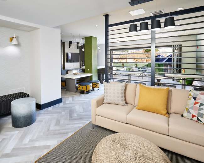 The image shows a modern lounge area with a beige sofa, geometric patterned cushions, and a view of a connected kitchen space.