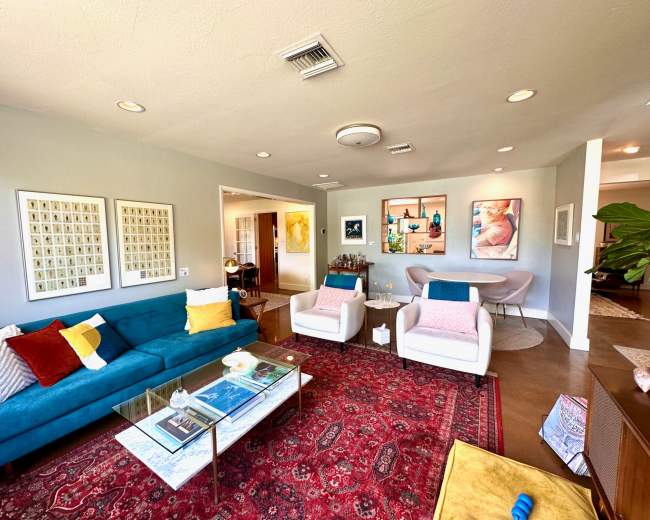 The living room features a blue sofa, two white armchairs, a glass coffee table, and a large area rug, with artwork and decorative items on the walls and surfaces.