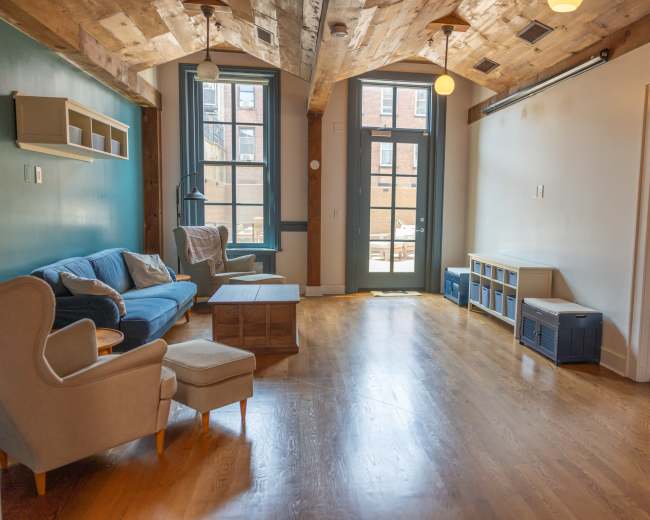 The image shows a spacious, well-lit living room with hardwood floors, featuring a blue sofa, a coffee table, and large windows that open to a patio area.