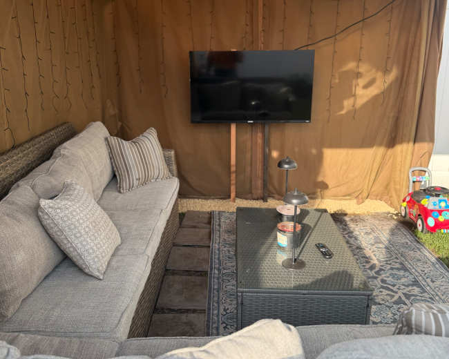 The image shows a cozy outdoor seating area with a light-colored woven sofa, a coffee table, and a television mounted on the wall inside a tent-like structure, surrounded by soft lighting.