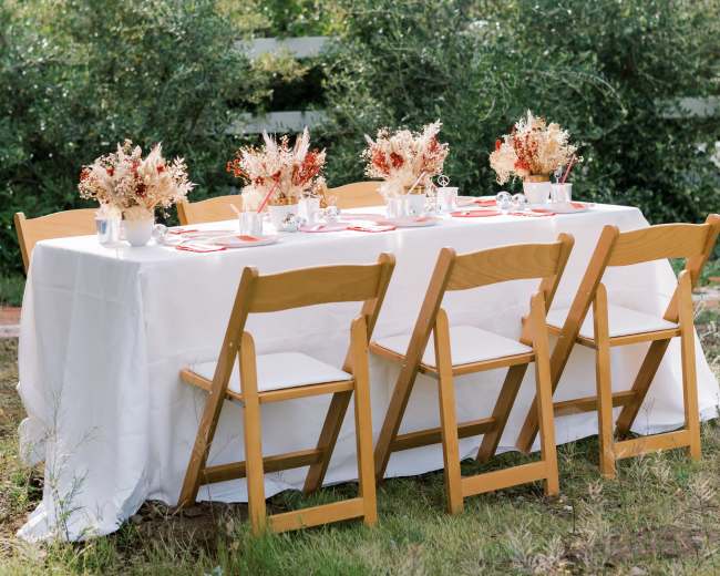 A long table set with floral arrangements and white tableware is arranged in a grassy outdoor area with wooden chairs around it.