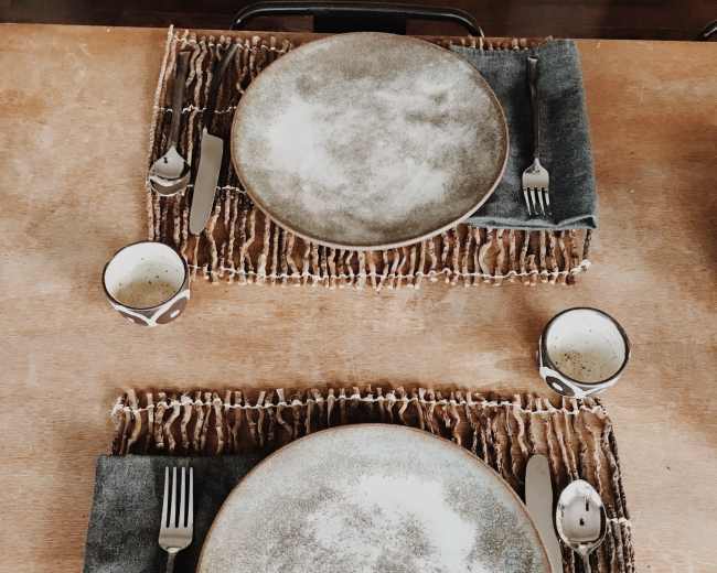 The image shows a dining table set with two plates, utensils, and cups arranged on woven mats.