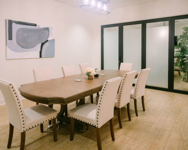 A long wooden dining table surrounded by eight upholstered chairs is placed in a well-lit room with a painting and glass doors leading to another area.