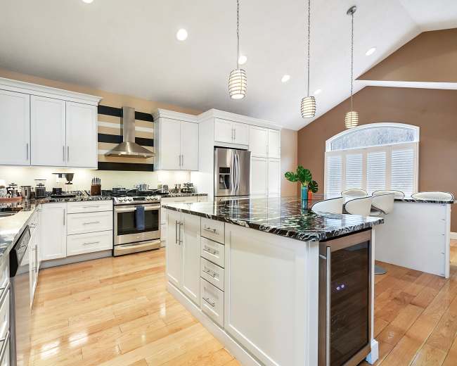 A modern kitchen features white cabinetry, a large island with dark granite countertops, stainless steel appliances, and pendant lighting.