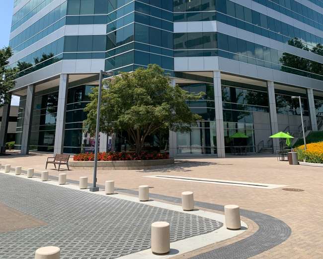 The image shows a modern glass office building with multiple stories, surrounded by landscaping and a paved walkway.