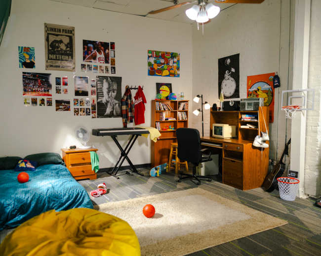 The image shows a cozy bedroom with a bed, a desk with a computer, a keyboard, and various posters on the walls.