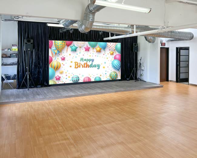 The image shows a spacious event room with a large "Happy Birthday" display on a screen, surrounded by colorful balloons and decorations.