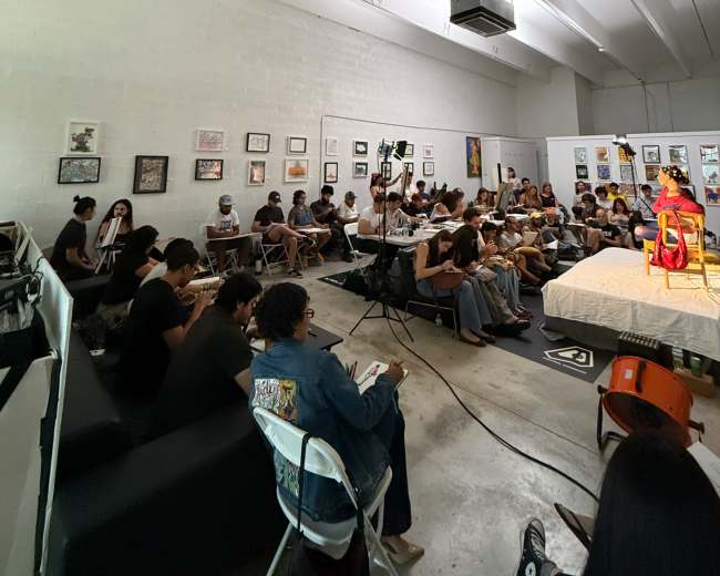 A diverse group of people sits attentively in an art space, facing a speaker on a stage surrounded by various artworks on the walls.