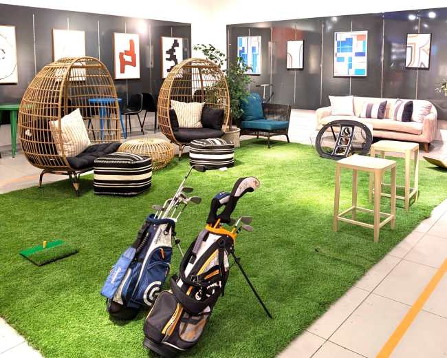 The image shows a modern indoor space featuring golf clubs, a green artificial turf area, stylish seating arrangements, and various artwork displayed on the walls.