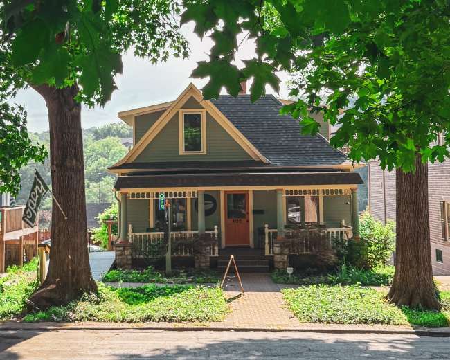 A green, two-story house with a front porch and large trees on either side is situated along a quiet street.