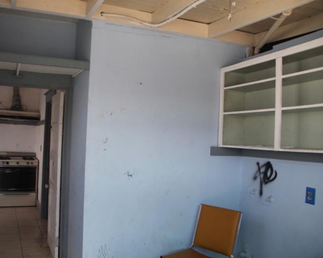 The image shows a small room with a blue wall, a chair, and a door leading to a kitchen area that includes a stove.