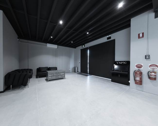 The image shows a minimalist interior space with a black couch, a metal coffee table, and a wall-mounted vending machine near a fire extinguisher.