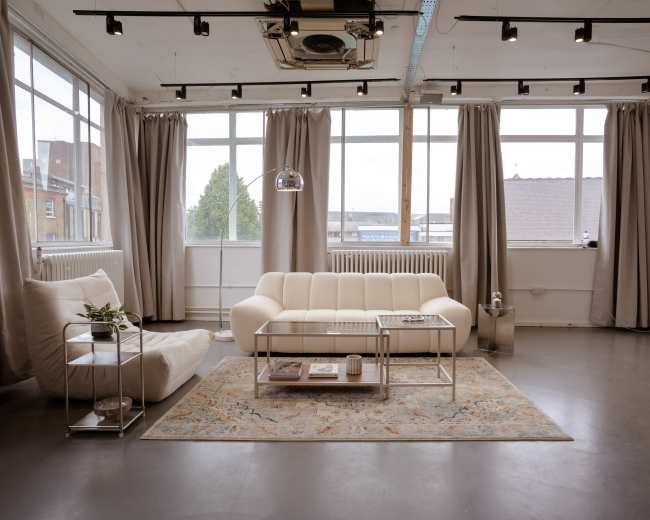 The image shows a modern, minimalist living room with large windows, a white sofa, a decorative area rug, and light curtains.