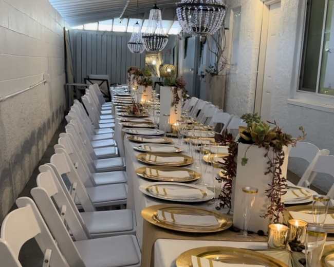 A long dining table is set with golden plates, glasses, and candles, surrounded by white chairs, under decorative chandeliers in a narrow indoor space.
