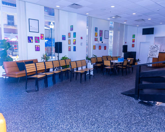 The image shows a spacious, modern interior with rows of wooden benches, a few potted plants, and colorful artwork displayed on the walls.