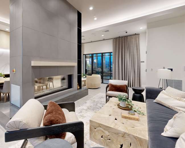 The image depicts a modern living space featuring a central fireplace, cozy seating areas, and large windows with natural light coming in.