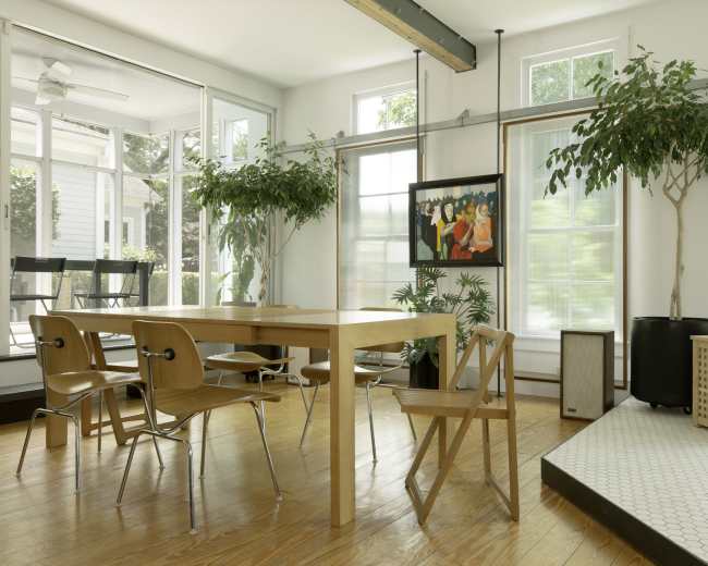 A well-lit dining area featuring a wooden table surrounded by chairs, with large windows, potted plants, and a painting on the wall.