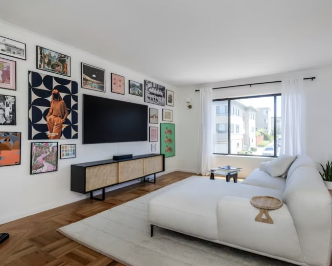 A light-filled living room features a large TV mounted on a sleek media unit, surrounded by a gallery of framed art on the walls and a view of the street beyond through a large window.