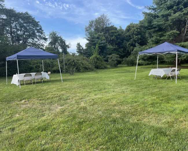 Two blue canopies are set up on a grassy area, each with tables and chairs underneath, surrounded by trees.